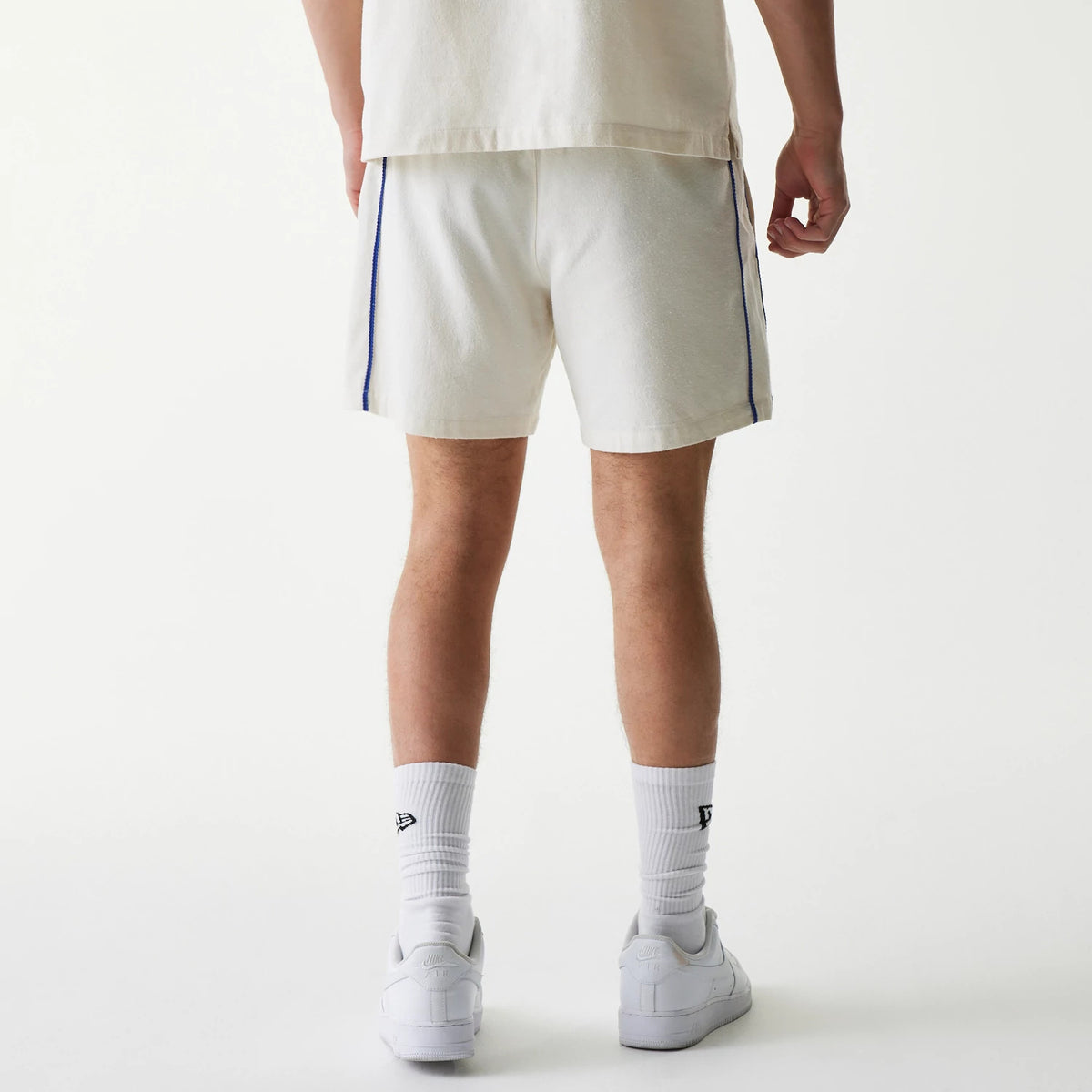 New Era New York Mets MLB Piping Open Cream Shorts, back view on-model showing the cream shorts with blue piping along the sides, paired with white socks featuring an embroidered New Era logo, styled with white trainers.