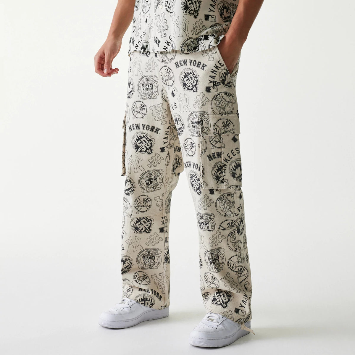 New Era MLB Subway Series All Over Print Cargo Trousers, front view on a model showing the intricate black and white Subway Series graphics with team logos, side cargo pockets, and a relaxed fit paired with white trainers.