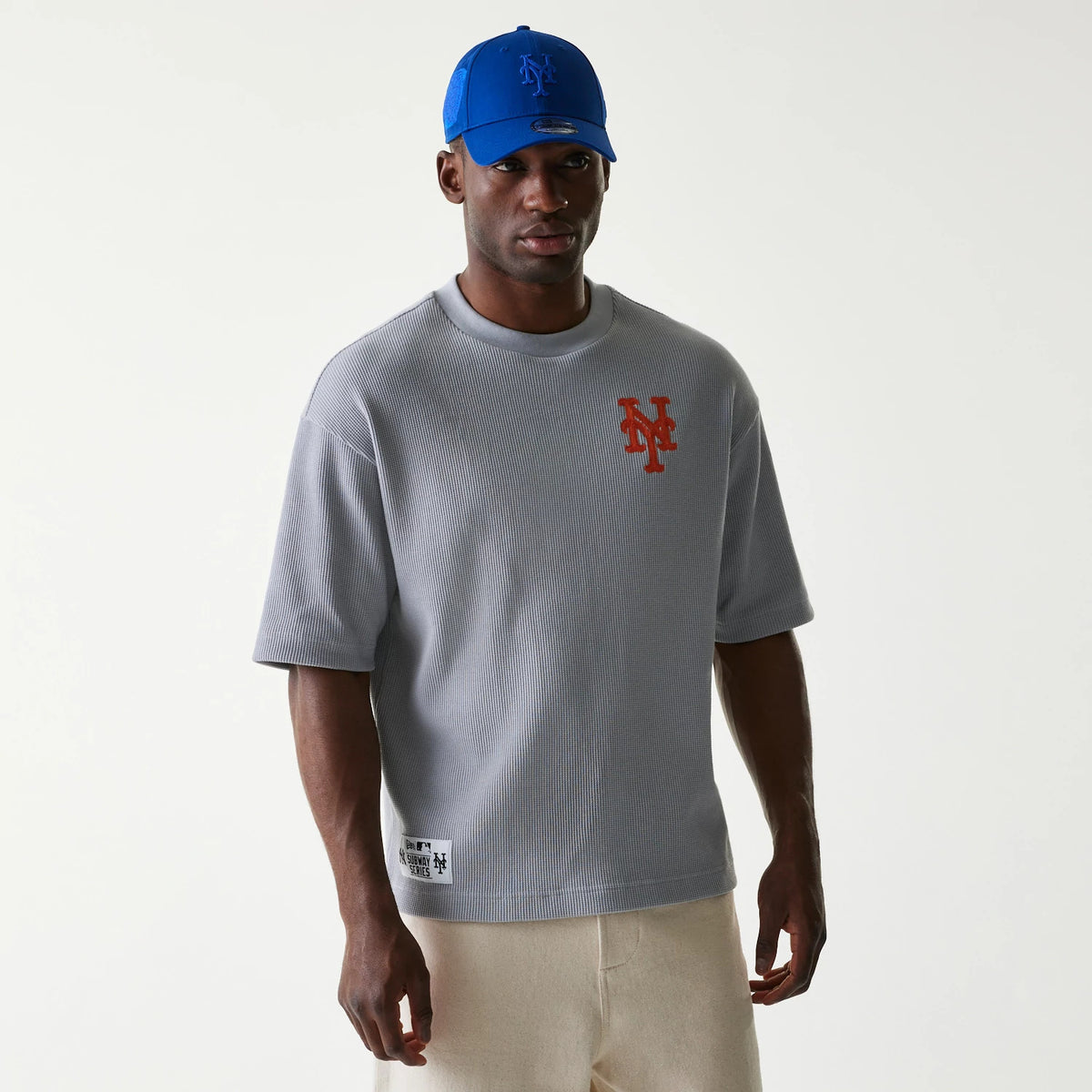 New Era New York Mets MLB Waffle Grey Oversized T-Shirt, front view on-model showing the orange embroidered Mets logo on the left chest, with a ribbed texture and a New Era patch near the bottom hem, styled with a blue cap.