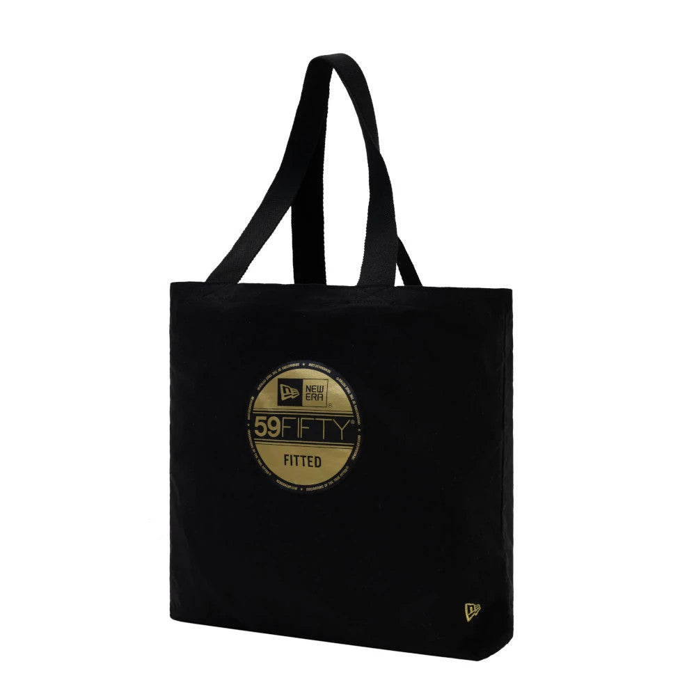 New Era Black Tote Bag, three-quarter view showing the prominent round gold 59FIFTY graphic printed on the front, long black handles for carrying, and a small embroidered New Era logo near the bottom corner.