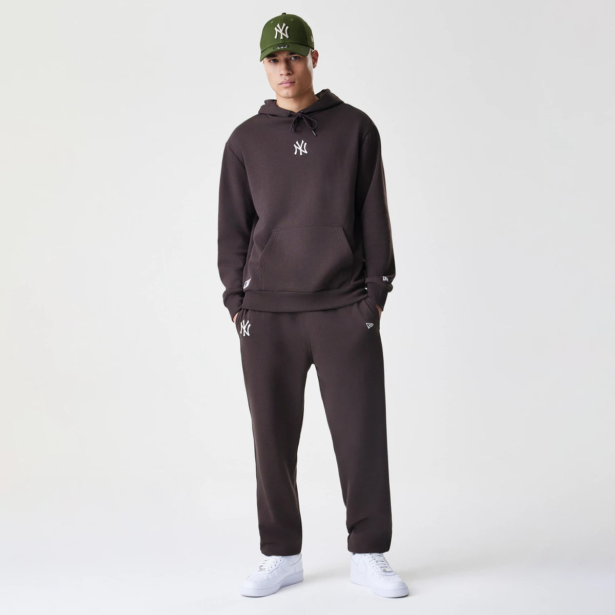 New Era New York Yankees MLB Midi League Essential Dark Brown Oversized Pullover Hoodie, front view on-model showing the embroidered white Yankees logo on the chest and left wrist, styled with matching dark brown joggers and a green cap.