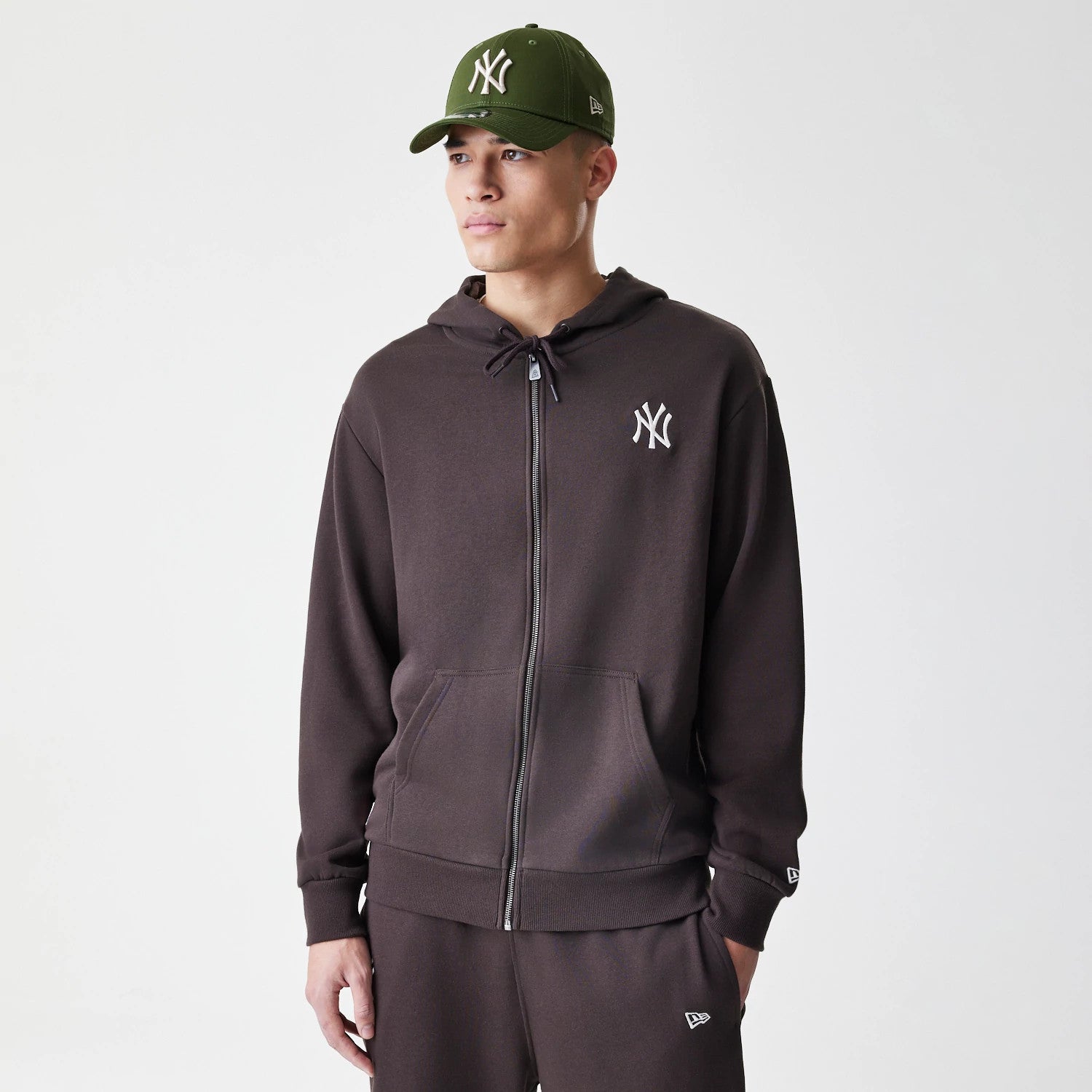 New Era New York Yankees MLB Midi League Essential Dark Brown Full Zip Hoodie, front view on-model showing the white embroidered Yankees logo on the left chest, with a full zip closure down the centre, paired with matching dark brown trousers.