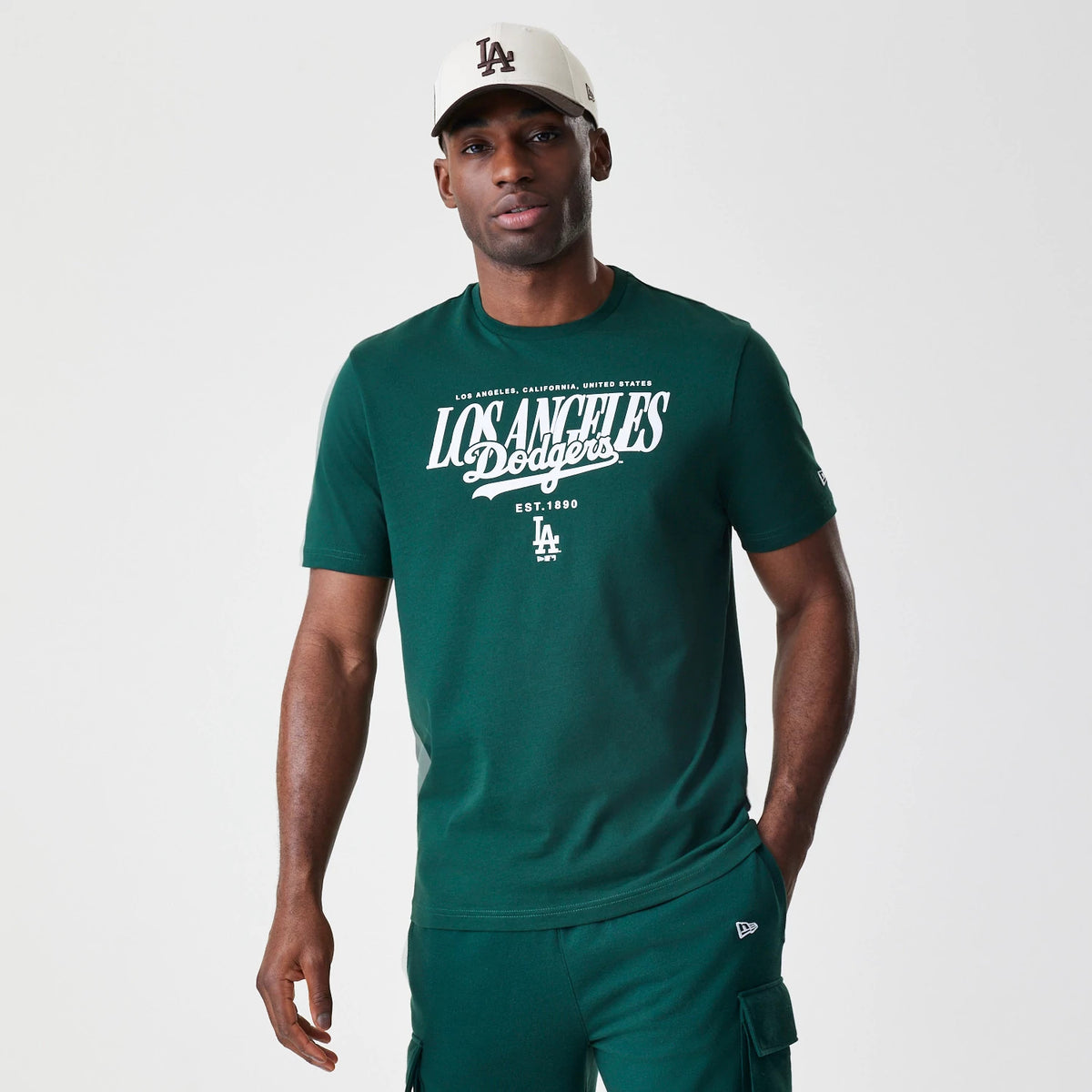 New Era LA Dodgers MLB Script Graphic Dark Green T-Shirt, front view on a model with a white cap, featuring Los Angeles Dodgers in large white script across the chest and an embroidered New Era logo on the sleeve.