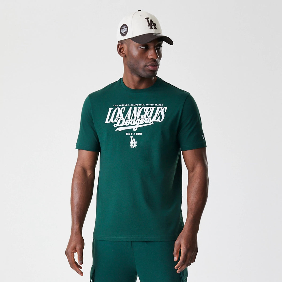 New Era LA Dodgers MLB Script Graphic Dark Green T-Shirt, front view on-model showing the white printed Los Angeles Dodgers script and LA logo on the chest with a small New Era logo on the left sleeve, paired with a contrasting cap.