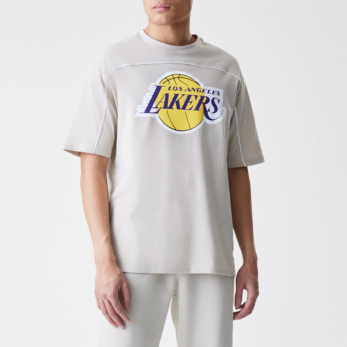 New Era LA Lakers NBA Piping Light Beige Oversized T-Shirt, front view on a model with matching trousers, featuring a large yellow basketball graphic and an embroidered Lakers logo across the chest with white piping.