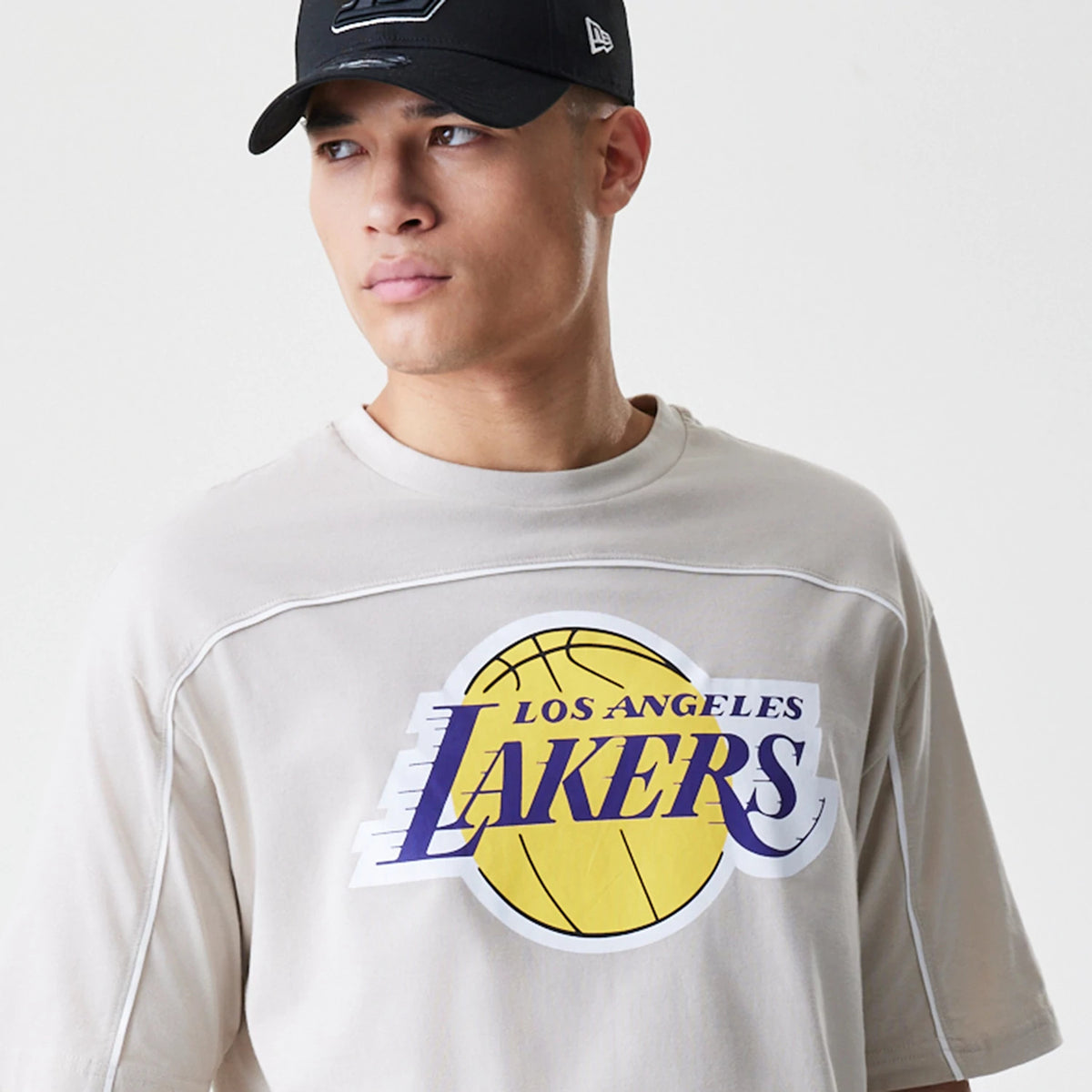 New Era LA Lakers NBA Piping Light Beige Oversized T-Shirt, front view on a model with a black cap, featuring a large Los Angeles Lakers graphic in yellow and purple across the chest and distinct piping along the seams.