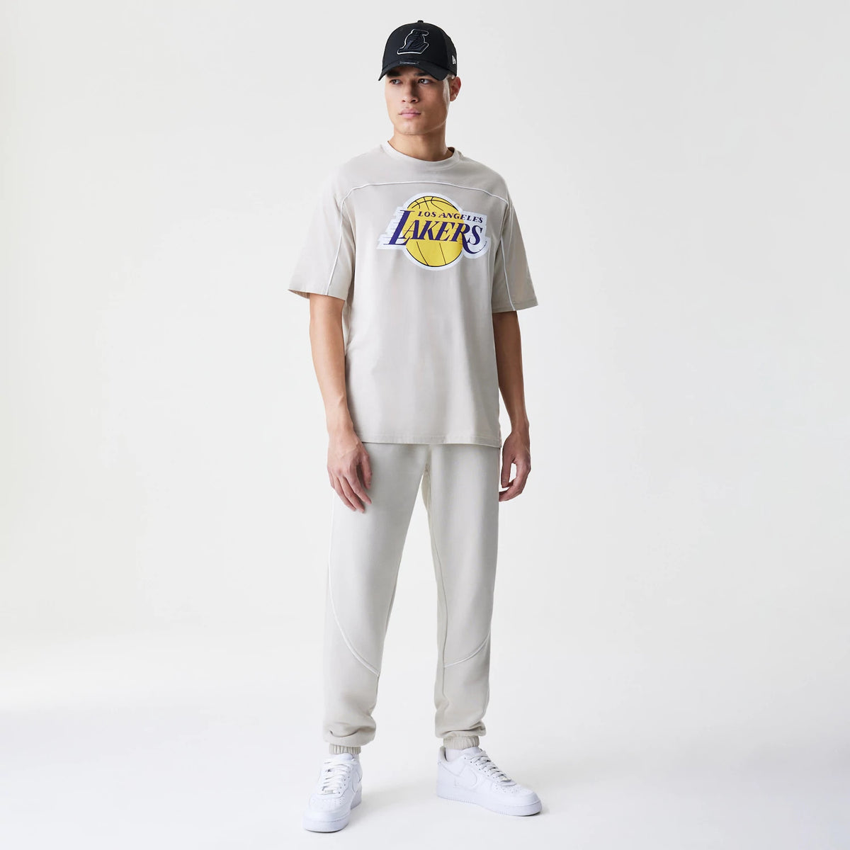 New Era LA Lakers NBA Piping Light Beige Oversized T-Shirt, front view on-model showing the purple and yellow Los Angeles Lakers logo printed across the chest with seam details on the shoulder piping, styled with matching light beige trousers.