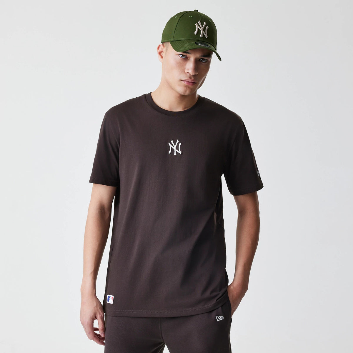New Era New York Yankees MLB Midi League Essential Dark Brown Oversized T-Shirt, front view on-model displaying the embroidered white Yankees logo centred on the chest, with short sleeves and a crew neck, styled with a matching green Yankees cap.