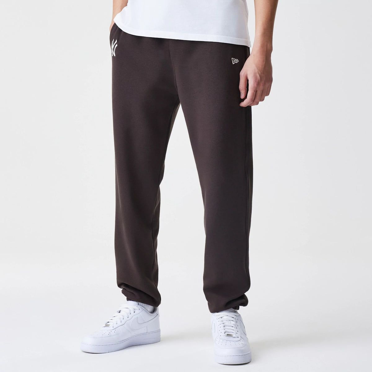New Era New York Yankees MLB Midi League Essential Dark Brown Joggers, front view on a model with a white embroidered Yankees logo on the left thigh and the New Era flag logo on the right thigh, paired with white trainers.