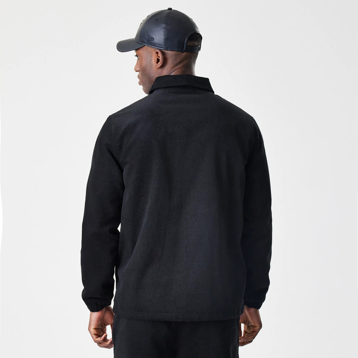 New Era Cord Black Coaches Jacket, back view on a model with matching black trousers and a cap, highlighting the plain black back and relaxed fit with a soft collar and long sleeves.