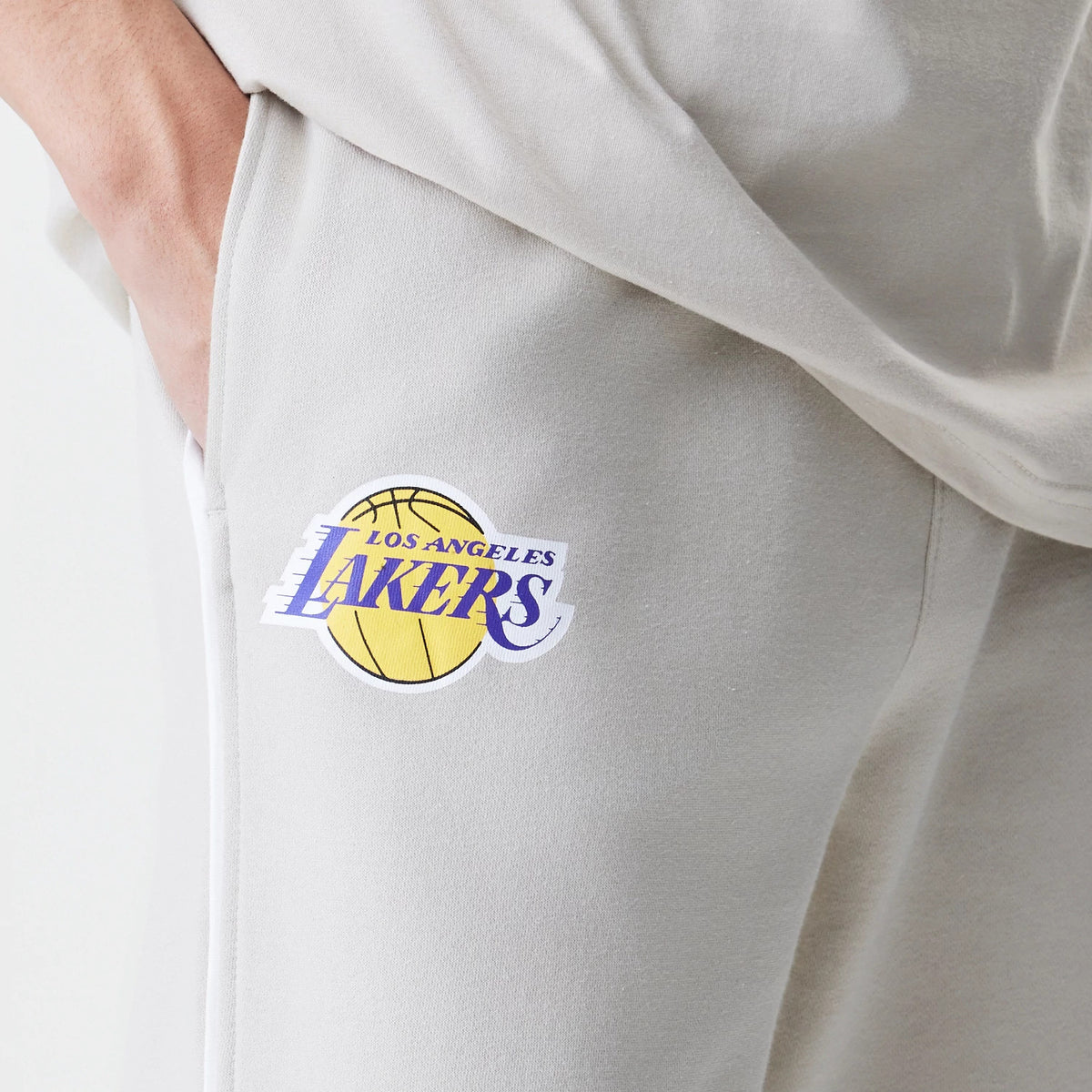 New Era LA Lakers NBA Piping Cream Joggers, close-up showing the Los Angeles Lakers purple and yellow basketball logo embroidered near the right pocket on a cream fabric, highlighting the soft texture and relaxed fit.