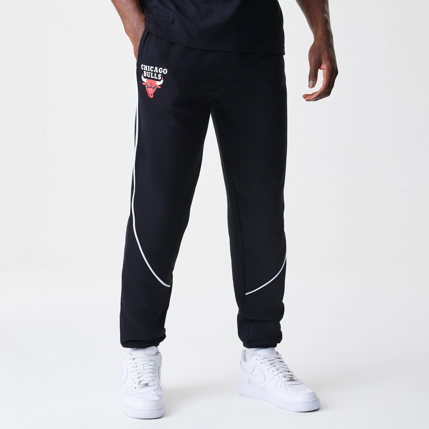 New Era Chicago Bulls NBA Piping Navy Joggers, front view on a model showing the red and white Bulls logo on the left thigh and contrasting white piping curving along the legs, paired with white trainers.