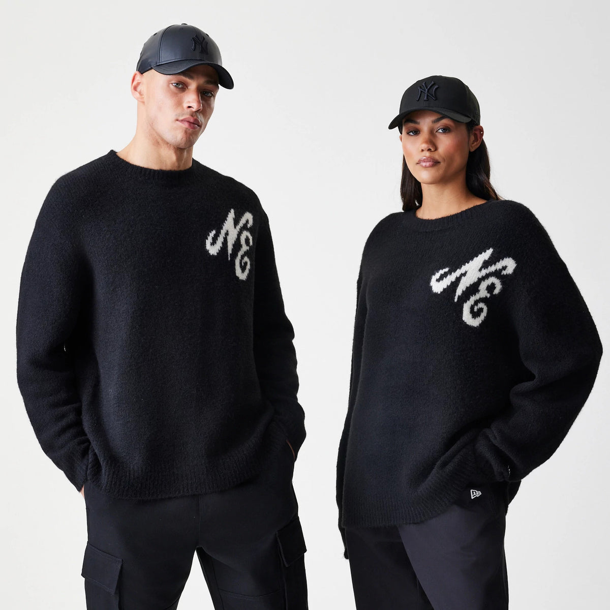New Era Knit Black Knitted Sweater, on-model view showing two models wearing the sweater with white cursive NE lettering on the left chest, paired with black trousers and black caps.