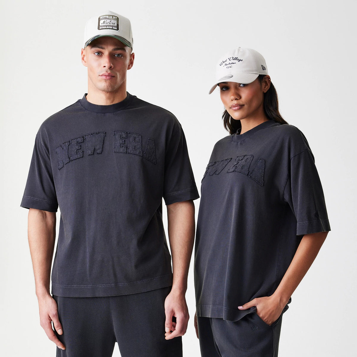 New Era Washed Black Oversized T-Shirt, front view on models showing an embroidered New Era logo across the chest of both shirts, styled with matching dark joggers and light-coloured caps, enhancing the relaxed fit and casual look.