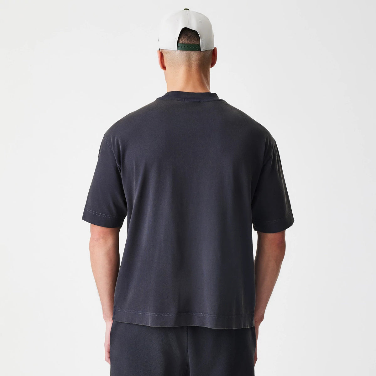 New Era Washed Black Oversized T-Shirt, back view showing the plain washed black fabric with no graphics or logos, relaxed fit, short sleeves, and straight hemline, styled with matching black trousers.