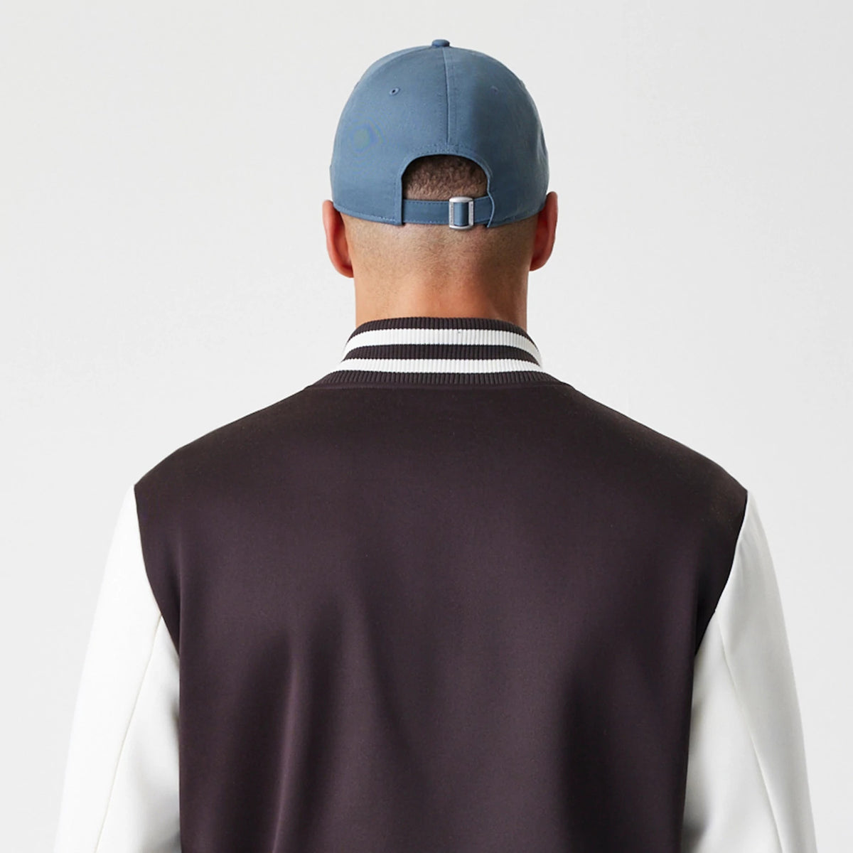 New Era New York Yankees MLB Dark Brown Varsity Jacket, back view on a model with a striped ribbed collar, contrasting white sleeves, and a dark brown back panel, styled with a light blue cap.