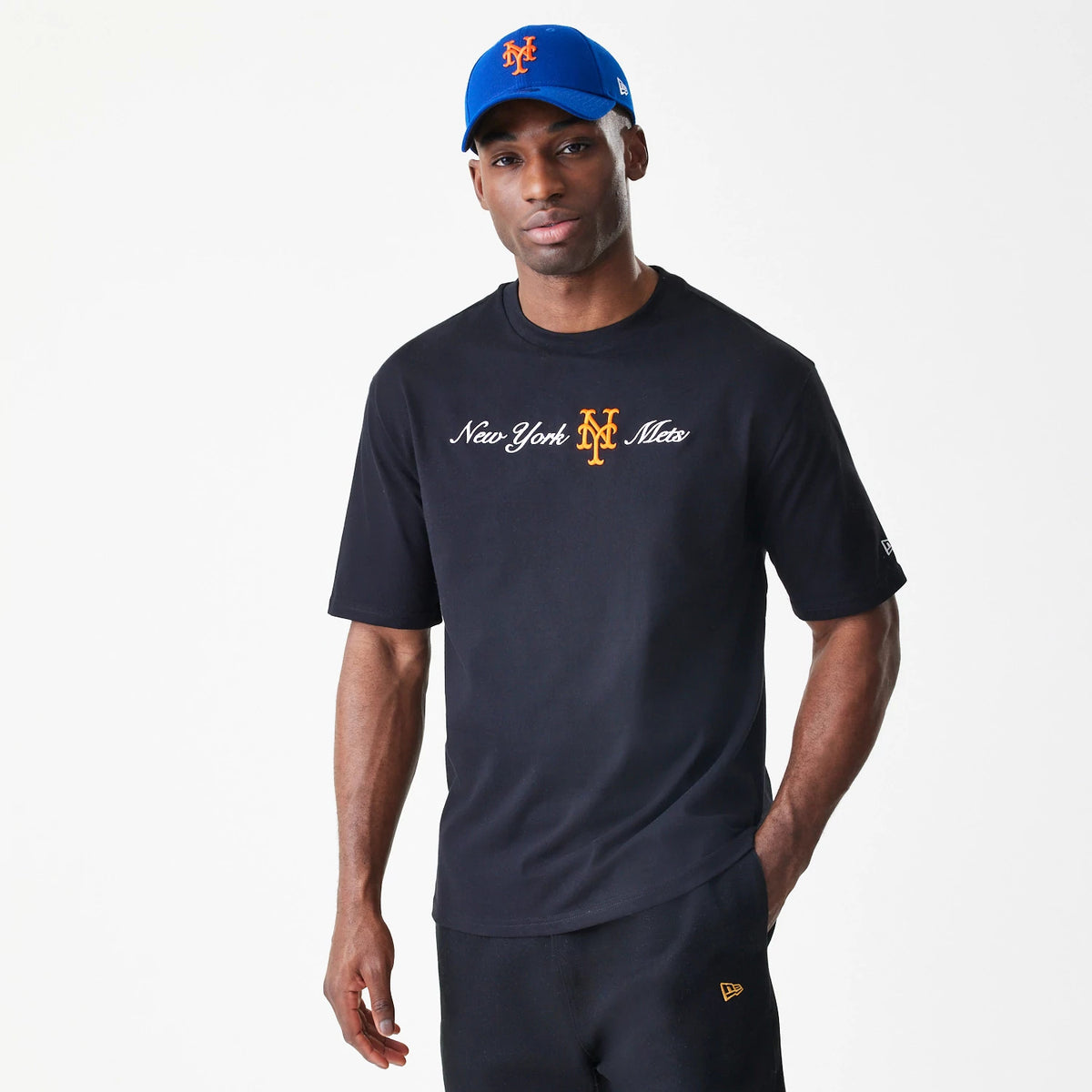 New Era New York Mets MLB Script Black Oversized T-Shirt, front view on-model showing white script lettering with an orange embroidered Mets logo on the chest, styled with black trousers featuring a yellow New Era logo near the pocket.