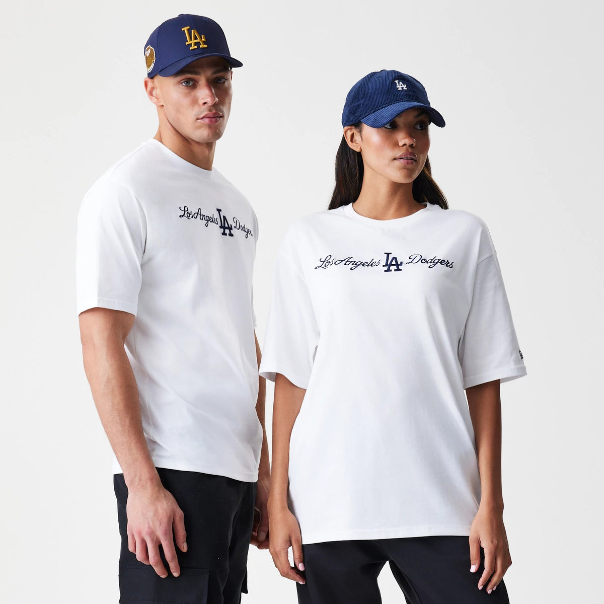 New Era LA Dodgers MLB Script White Oversized T-Shirt, front view on-model showing Los Angeles Dodgers script embroidered in black across the chest and a small patch on the left sleeve, paired with navy caps featuring the LA logo.