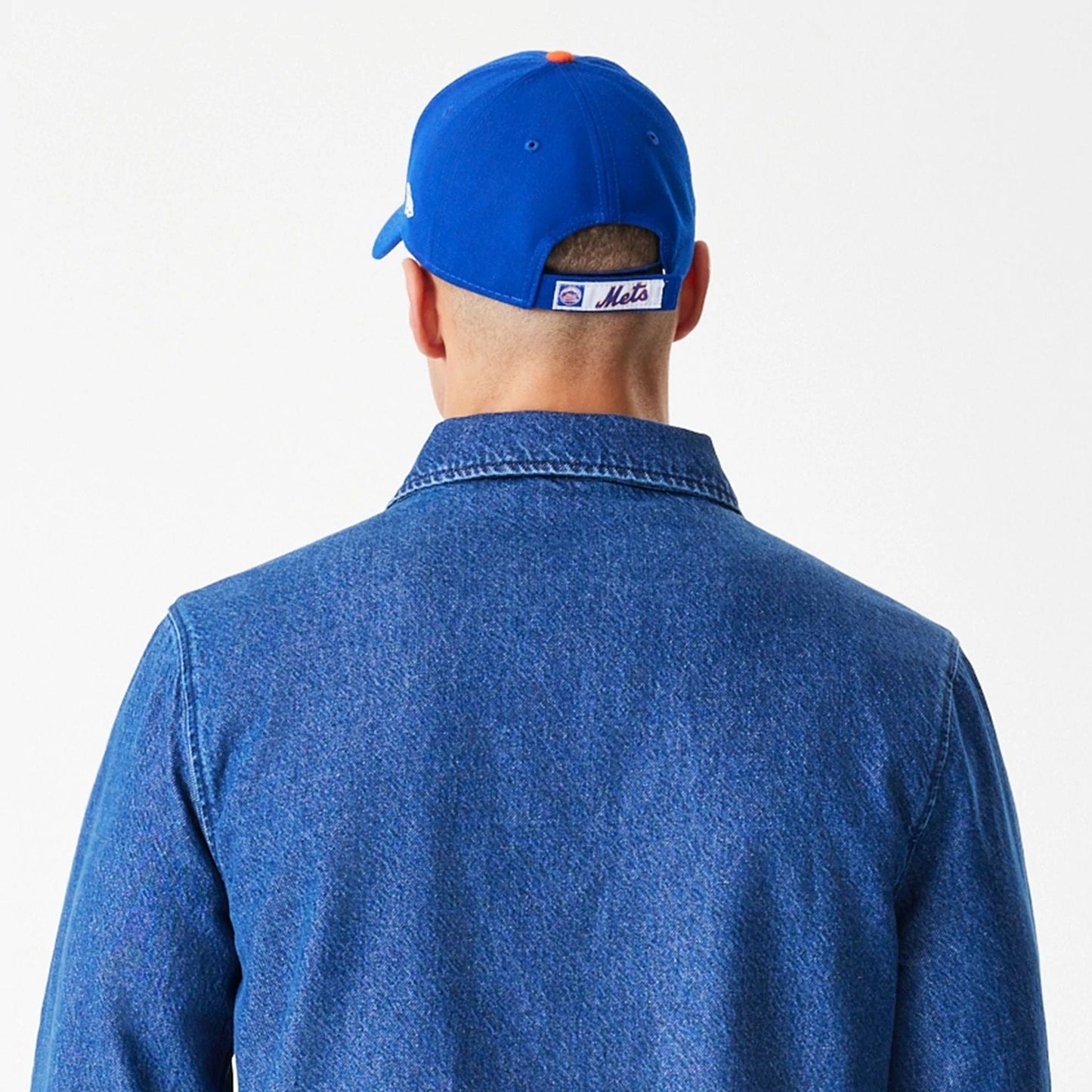 New Era New York Mets MLB Denim Dark Blue Coaches Jacket, back view on-model showing the plain dark blue denim with a collar at the neck, styled with a blue cap featuring a Mets logo label above the strap.