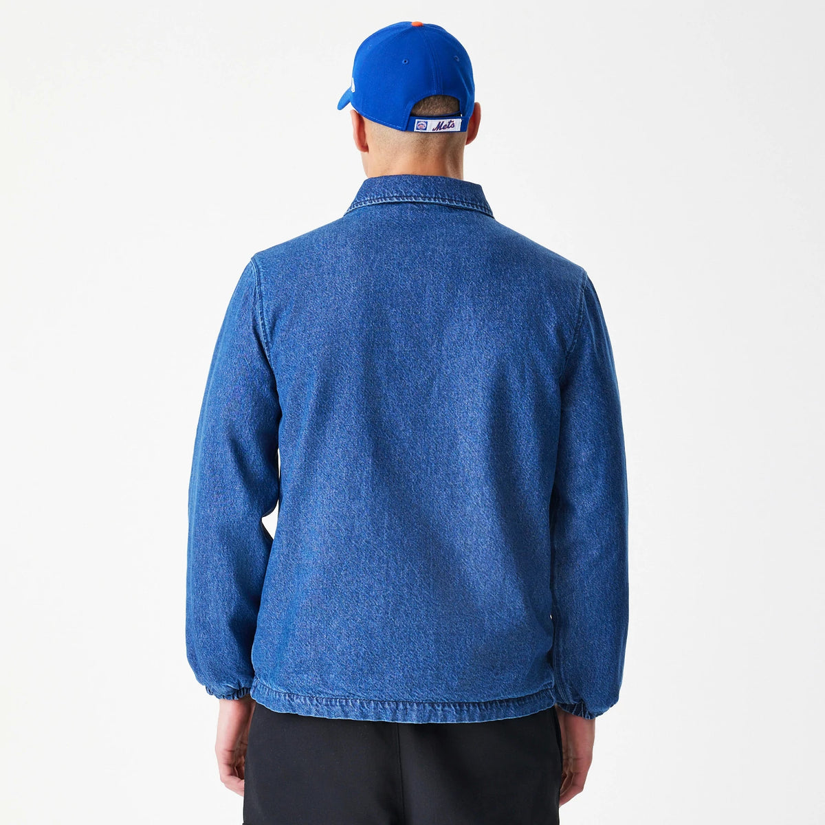 New Era New York Mets MLB Denim Dark Blue Coaches Jacket, back view on-model showcasing the plain dark blue denim with a relaxed collar and elastic cuffs, styled with a contrasting blue cap featuring Mets lettering above the adjustable strap.