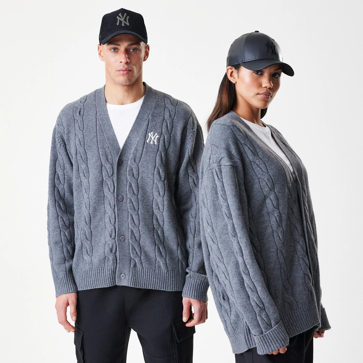 New Era New York Yankees MLB Cable Knit Dark Grey Cardigan, three-quarter view on models showing the prominent cable knit texture, ribbed cuffs and hem, with a white embroidered Yankees logo on the left chest, paired with black and grey caps.