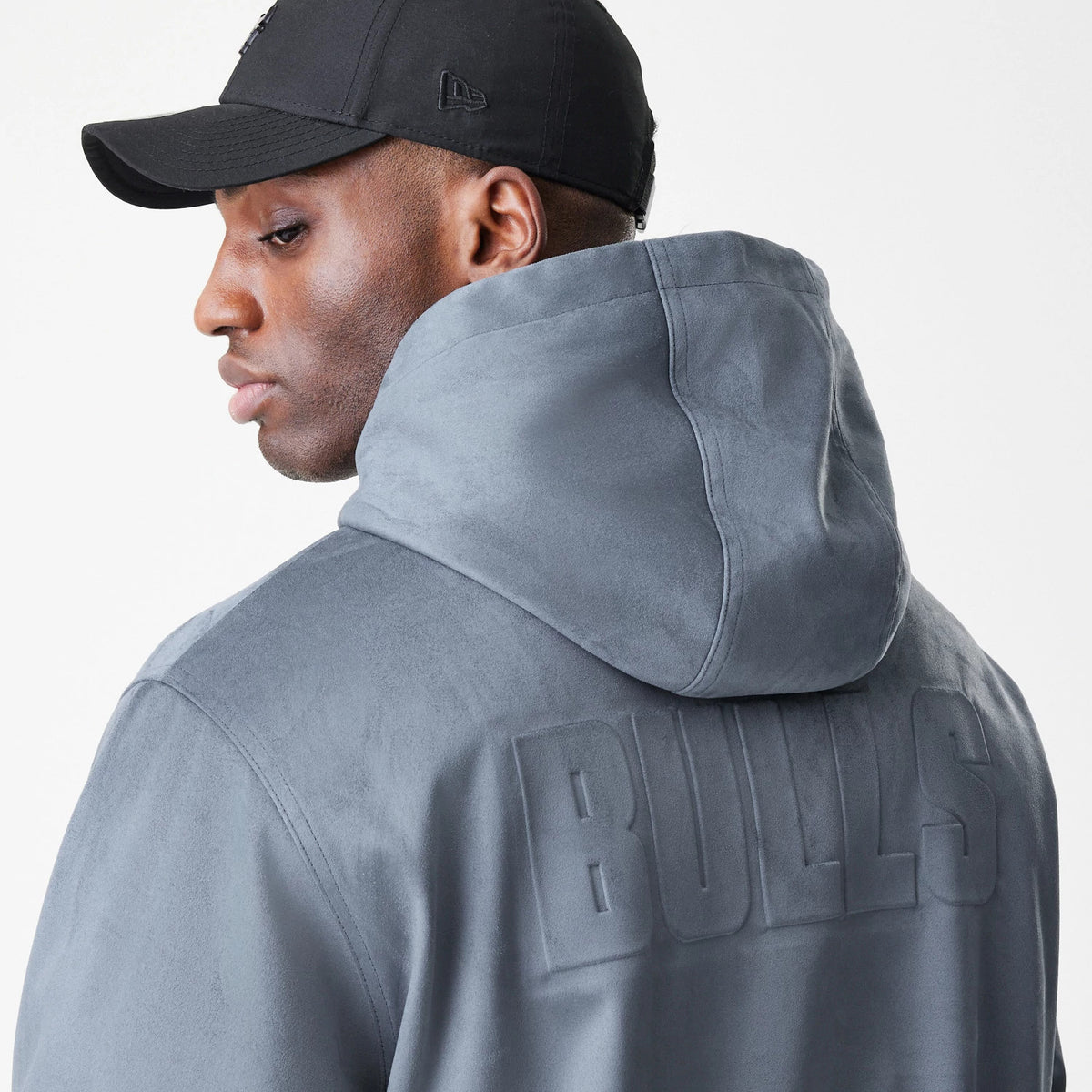 New Era Chicago Bulls NBA Suede Dark Grey Jacket, back view on a model featuring the raised BULLS lettering across the shoulders and a hood, worn with a black cap showing an embroidered New Era logo on the side.