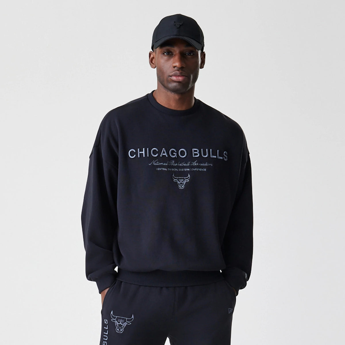 New Era Chicago Bulls NBA Linear Black Crew Neck Sweater, front view on a model, featuring grey Chicago Bulls text across the chest with additional lettering below and a small bull logo, set on a loose-fit black fabric.