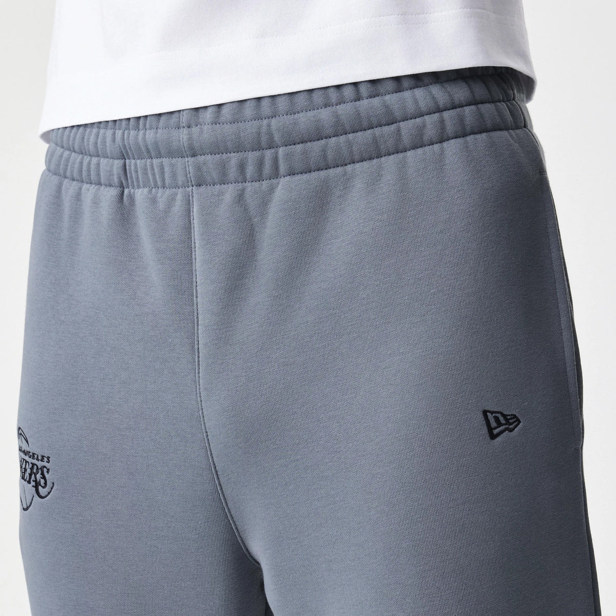 New Era LA Lakers NBA Linear Dark Grey Straight Leg Joggers, side view showing the embroidered New Era flag logo on the right thigh and the LA Lakers logo on the left, featuring a ribbed waistband with stitching.