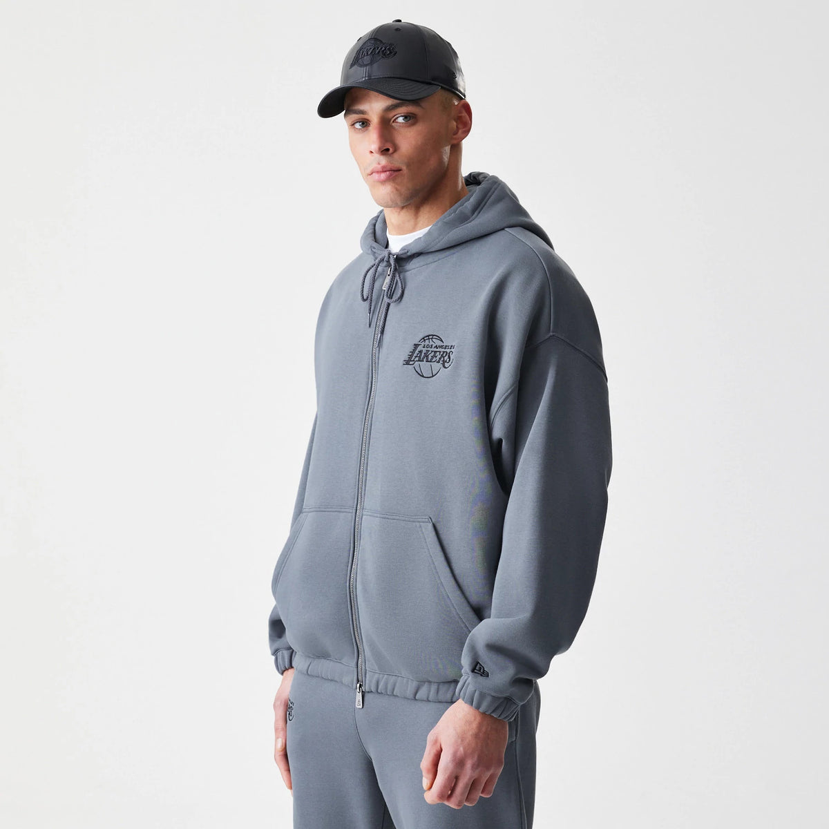 New Era LA Lakers NBA Linear Dark Grey Oversized Full Zip Hoodie, three-quarter view on-model showing the dark grey hoodie with a raised LA Lakers logo on the left chest, full zip closure, and a New Era logo on the left cuff.