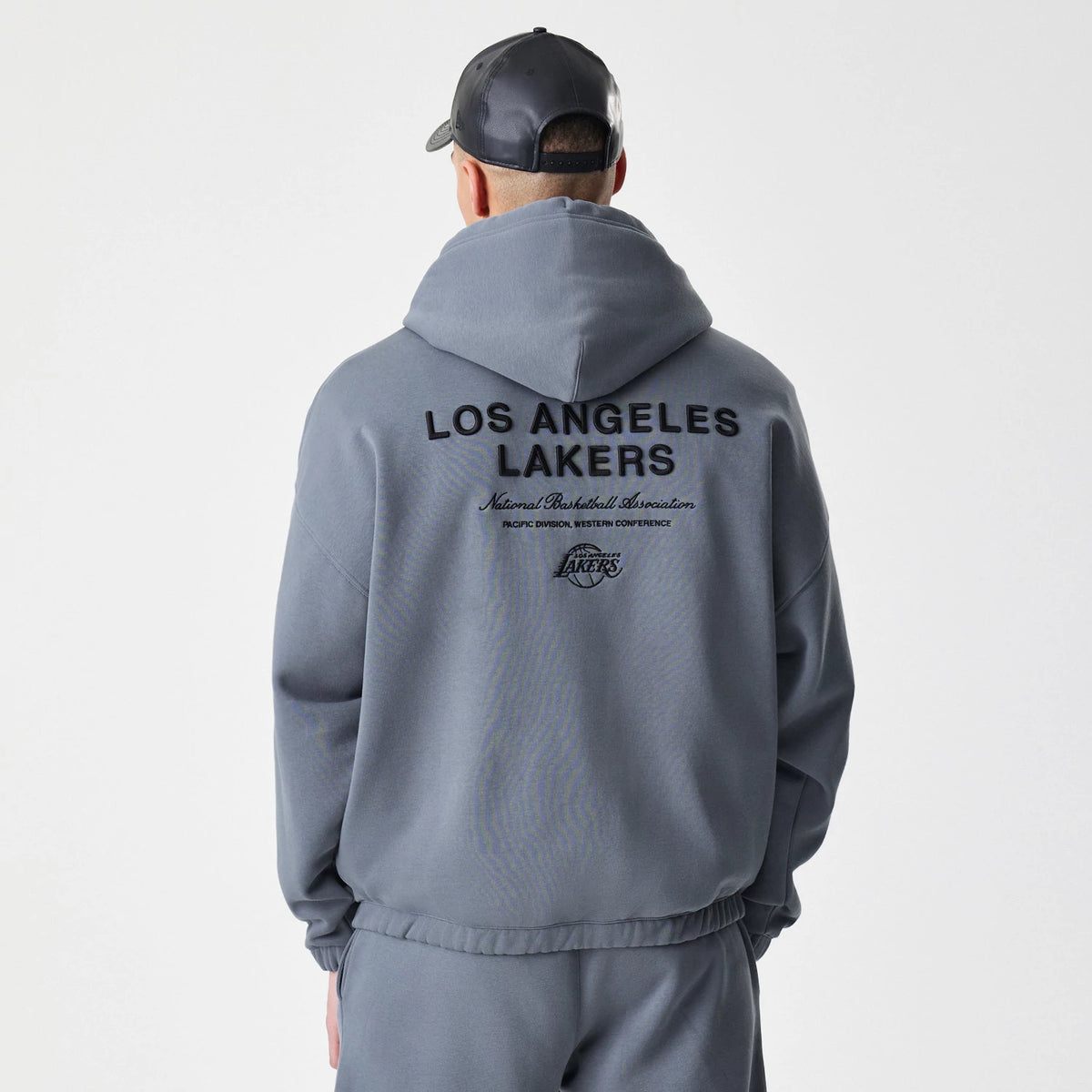 New Era LA Lakers NBA Linear Dark Grey Oversized Full Zip Hoodie, back view showcasing black printed Los Angeles Lakers text and graphics across the shoulders, worn by a model with matching dark grey trousers and a cap.