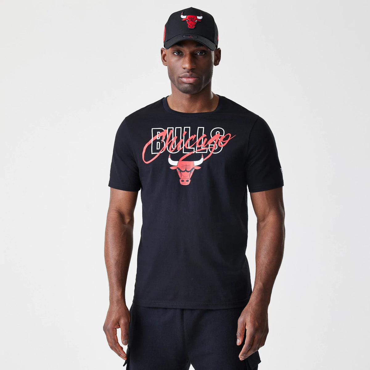 New Era Chicago Bulls NBA Script Black T-Shirt, front view on-model showing the red Bulls lettering with a script-style Chicago overlay, large bull logo beneath on the chest, paired with a black cap featuring an embroidered team logo.