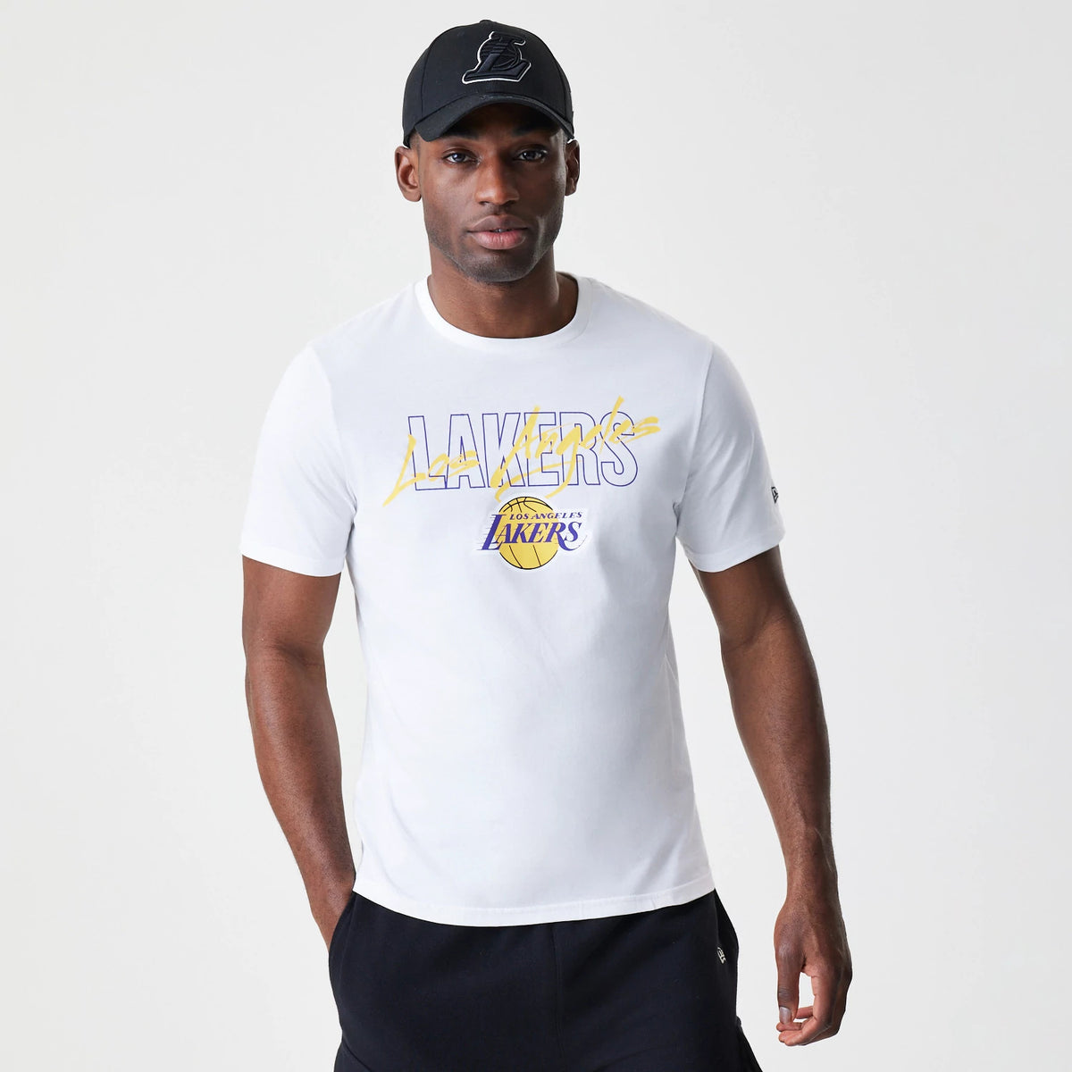 New Era LA Lakers NBA Script White T-Shirt, front view on-model showing the Lakers script graphic across the chest in yellow and purple with a small Los Angeles Lakers logo beneath, paired with a black cap.