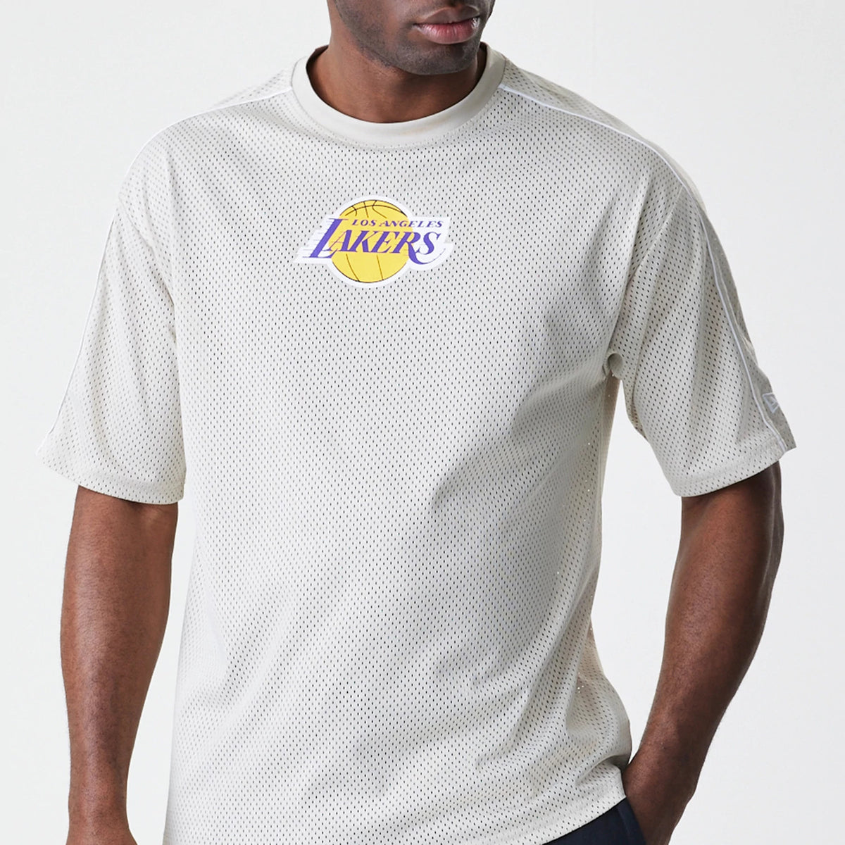 New Era LA Lakers NBA Piping Light Beige Oversized Mesh T-Shirt, front view showing the prominent Los Angeles Lakers embroidered logo in purple and yellow at the centre of the chest, worn by a model styled with dark trousers.