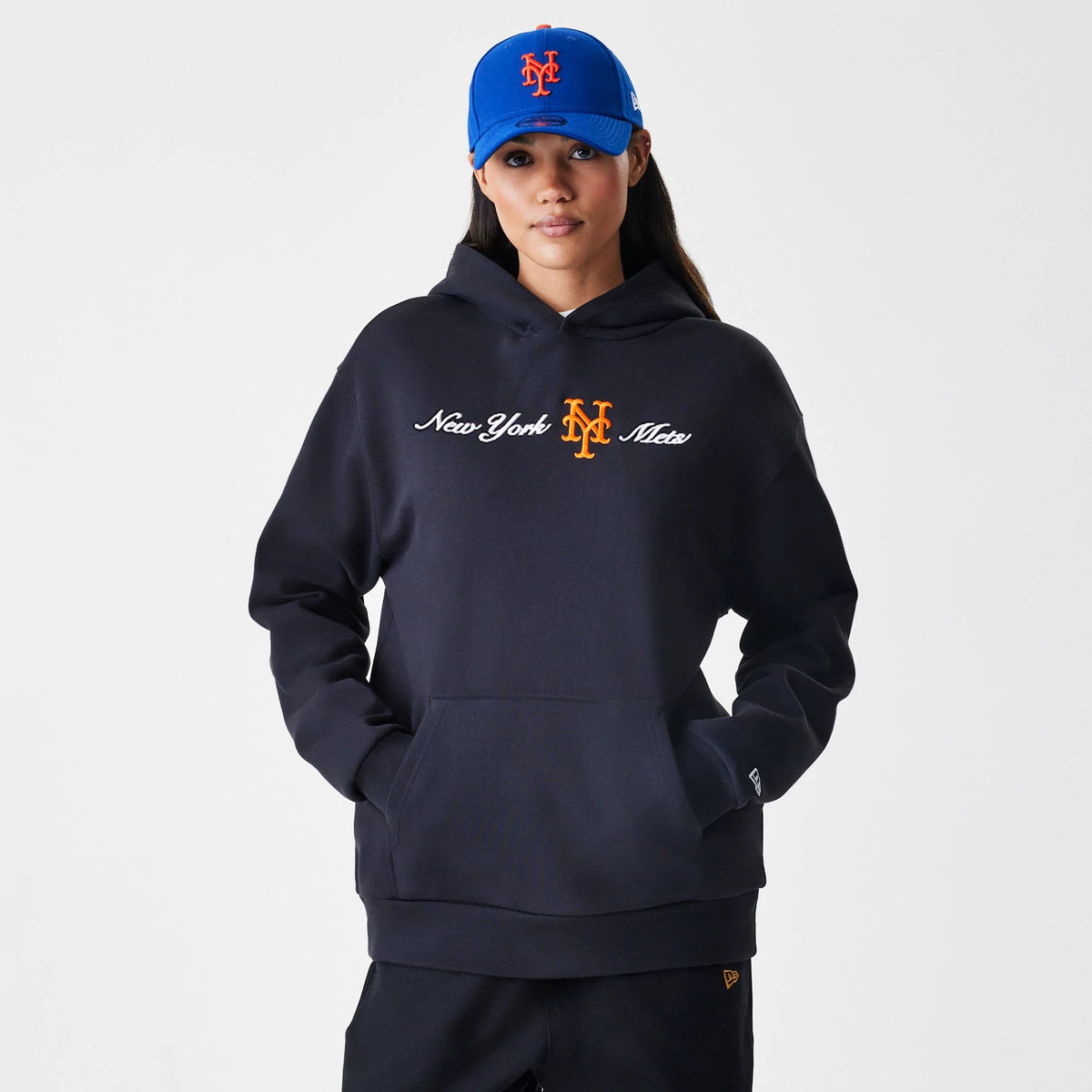 New Era New York Mets MLB Script Black Oversized Pullover Hoodie, front view on a model wearing a matching blue cap, featuring New York Mets embroidered in white and orange script across the chest and the New Era logo embroidered on the cuff.