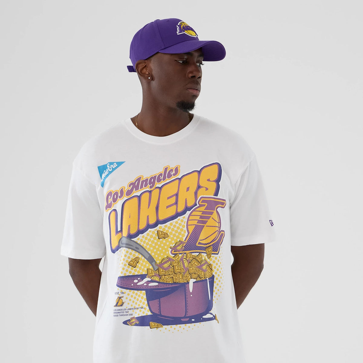 New Era LA Lakers NBA Lifestyle Graphic White T-Shirt, front view on a model with a purple cap showing a graphic of the Lakers' logo, a basketball theme in purple and yellow with Los Angeles Lakers text, and a New Era logo label on the sleeve.