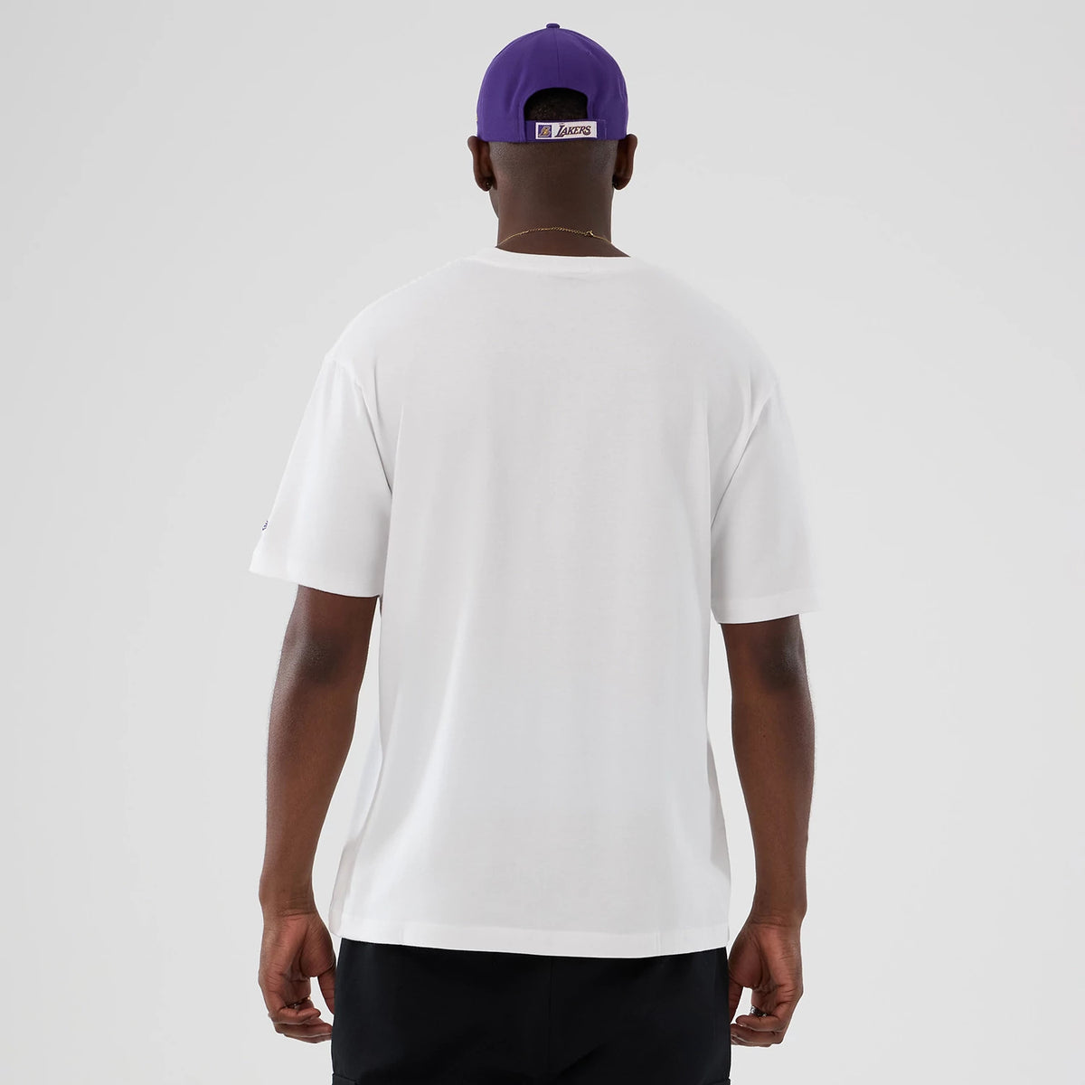 New Era LA Lakers NBA Lifestyle Graphic White T-Shirt, back view on a model showing the plain white surface with short sleeves, complemented by a purple cap with an embroidered LA Lakers logo on the back strap.