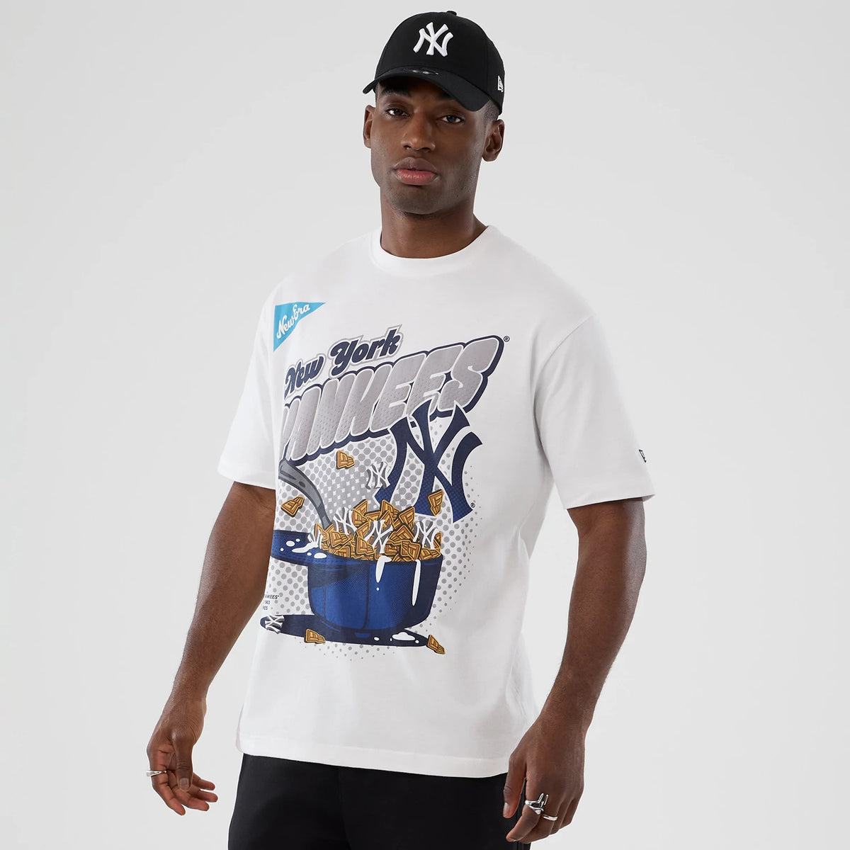 New Era New York Yankees MLB Lifestyle Graphic White T-Shirt, front view showing a graphic with New York Yankees lettering and a stylised bowl of cereal, worn by a model with a black cap featuring an embroidered white NY logo.