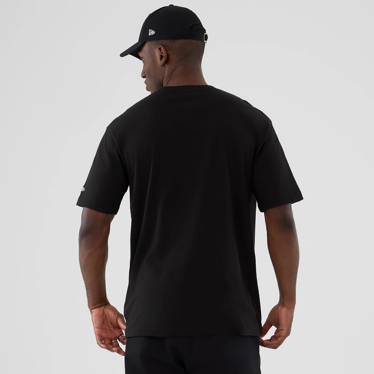 New Era National Lampoon's Christmas Vacation Lifestyle Graphic Black T-Shirt, back view on-model featuring plain black fabric with a small embroidered logo on the right sleeve and simple styling paired with a black cap.