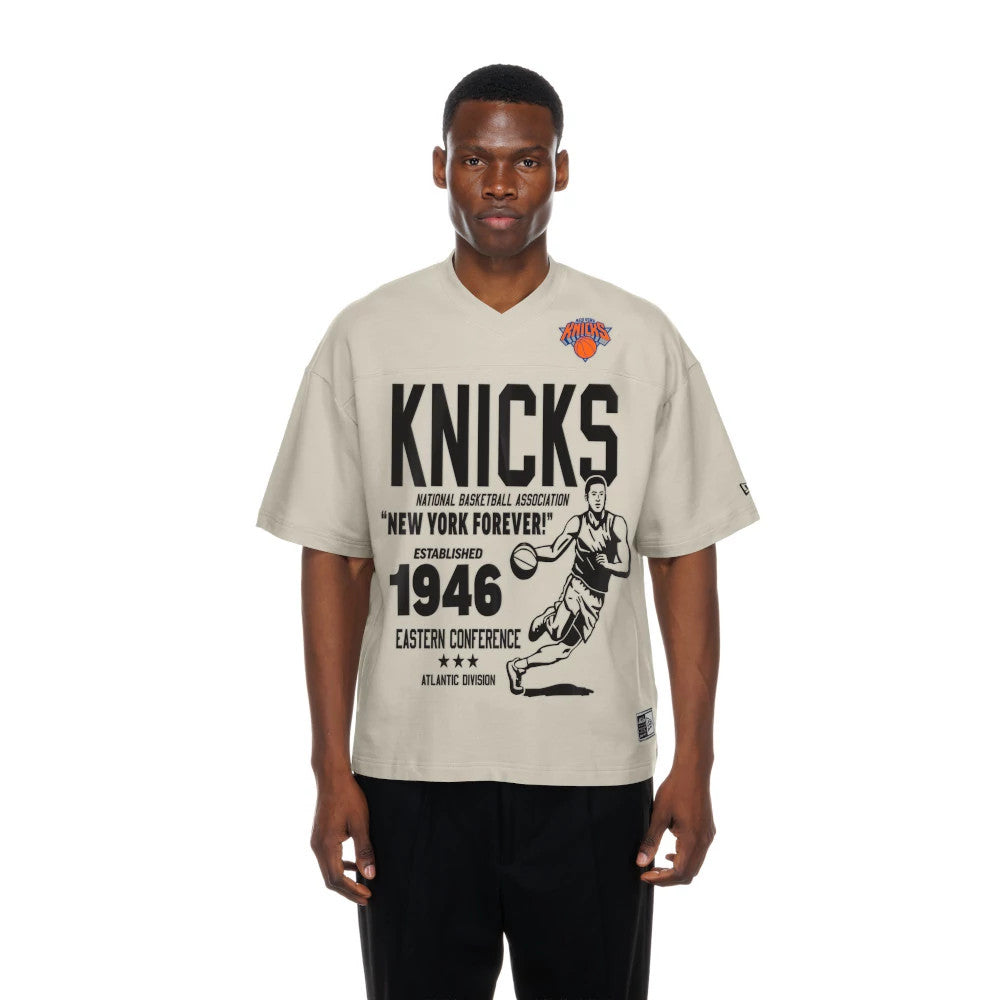 New Era New York Knicks NBA Throwback Prep Light Beige T-Shirt, front view showing black KNICKS lettering and vintage basketball graphics, with the orange and blue embroidered team logo on the left chest, model styled with black trousers.