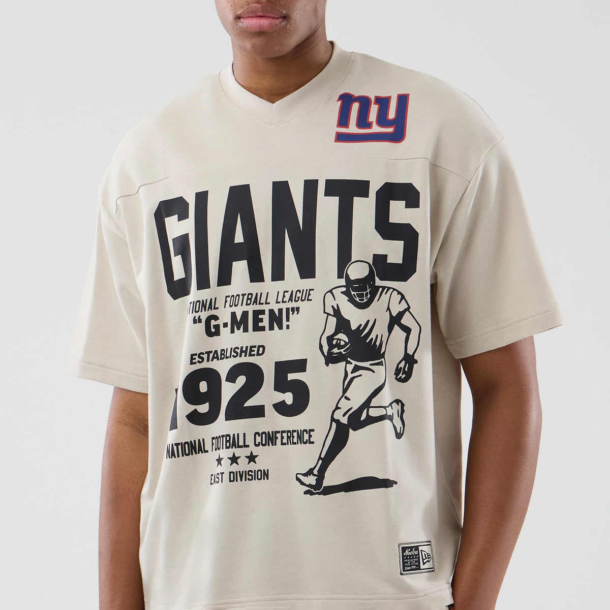 New Era New York Giants NFL Throwback Prep Light Beige T-Shirt, front view on a model showing black G-MEN lettering, a running football player graphic, the year 1925, an embroidered New Era logo patch near the hem, and an ny emblem on the chest.