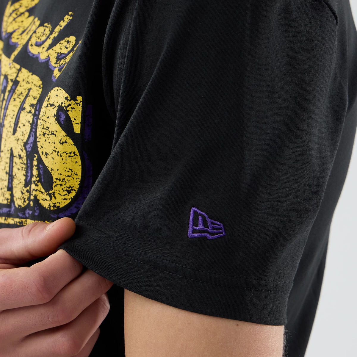 New Era LA Lakers NBA City Edition Black T-Shirt, close-up side view showing a purple embroidered New Era logo on the sleeve and the yellow and purple Lakers graphic partially on the chest.