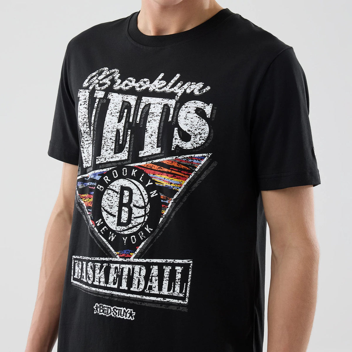 New Era Brooklyn Nets NBA City Edition Black T-Shirt, front view showing a graphic with distressed white Brooklyn Nets lettering, a stylised basketball logo, and colourful accents, worn by a model.