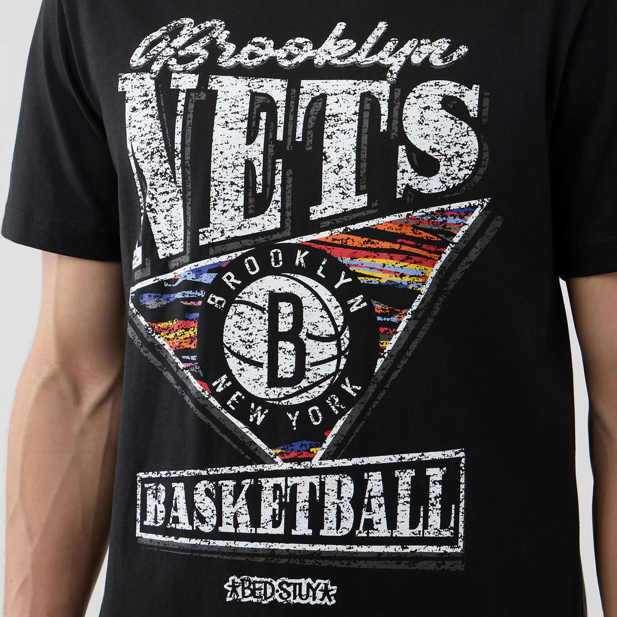 New Era Brooklyn Nets NBA City Edition Black T-Shirt, front view showing a large distressed Brooklyn Nets graphic with a multicoloured triangle design and a basketball motif at the centre, worn on a model.
