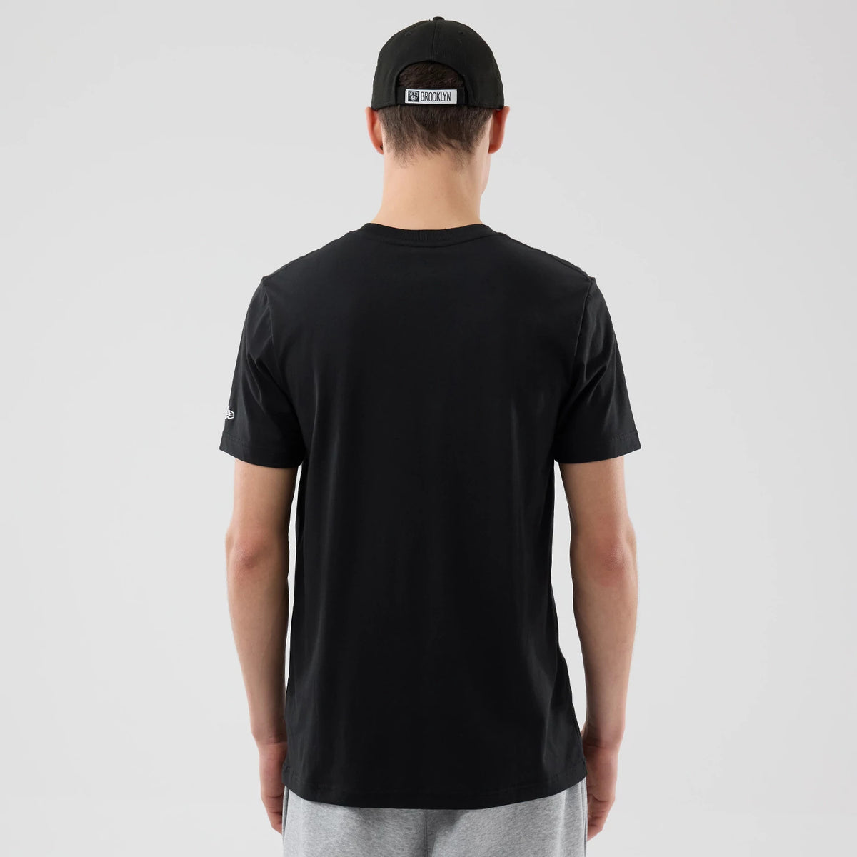 New Era Brooklyn Nets NBA City Edition Black T-Shirt, back view on-model showing the plain black fabric with a small embroidered logo on the sleeve, styled with a black cap featuring a white Brooklyn label displayed centrally above the strap.