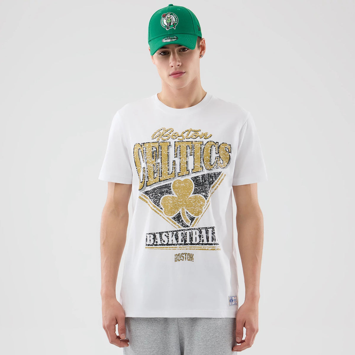 New Era Boston Celtics NBA City Edition White T-Shirt, front view on a model wearing a green cap, featuring Celtics lettering and graphics in gold and black on the chest with a small matching logo near the hemline.