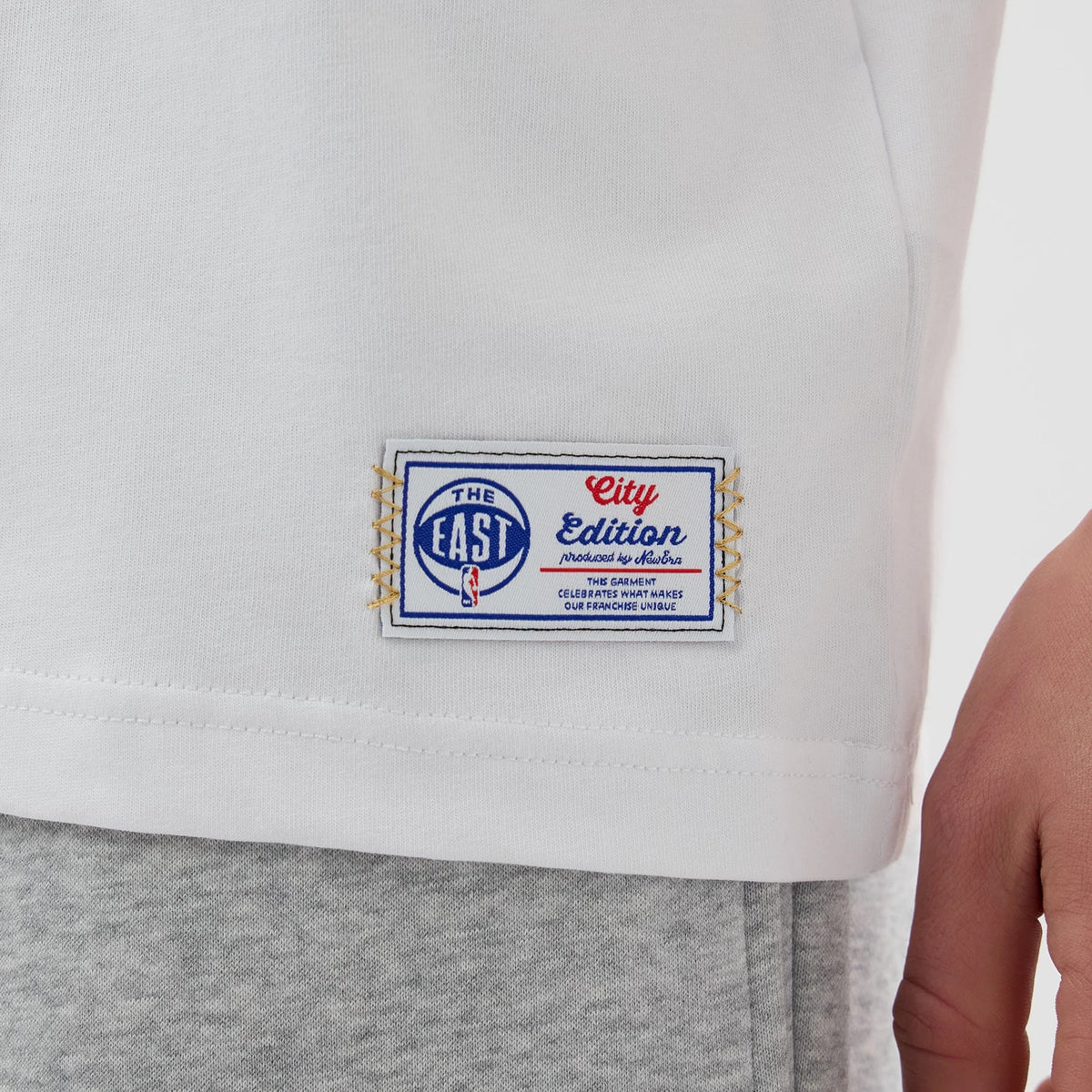New Era Boston Celtics NBA City Edition White T-Shirt, side view showing a detailed woven patch with The East and City Edition embroidered text above the NBA logo at the bottom edge of the shirt.