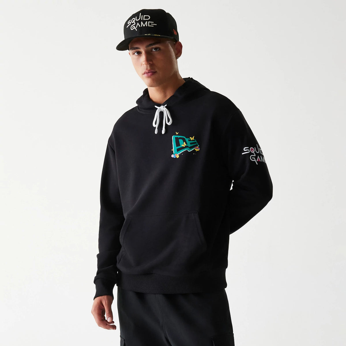 New Era Netflix Squid Game Black Oversized Pullover Hoodie, front view on-model featuring a colourful logo on the chest, white drawstrings at the hood, and Squid Game lettering embroidered on the sleeve.
