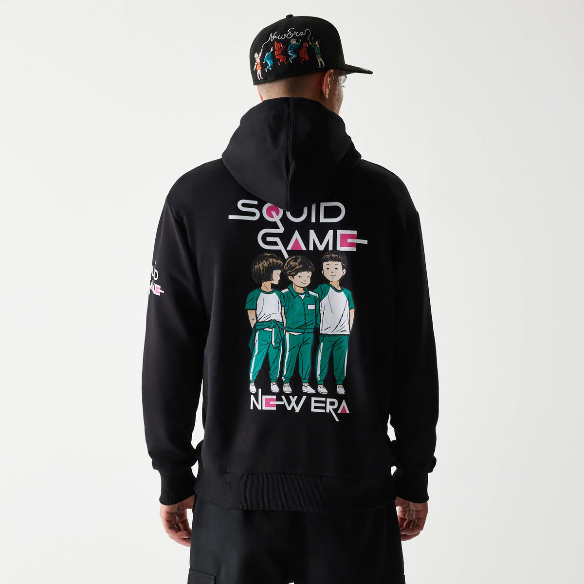 New Era Netflix Squid Game Black Oversized Pullover Hoodie, back view on a model wearing a black cap, showing a colourful graphic of three characters with Squid Game and New Era lettering across the upper back.