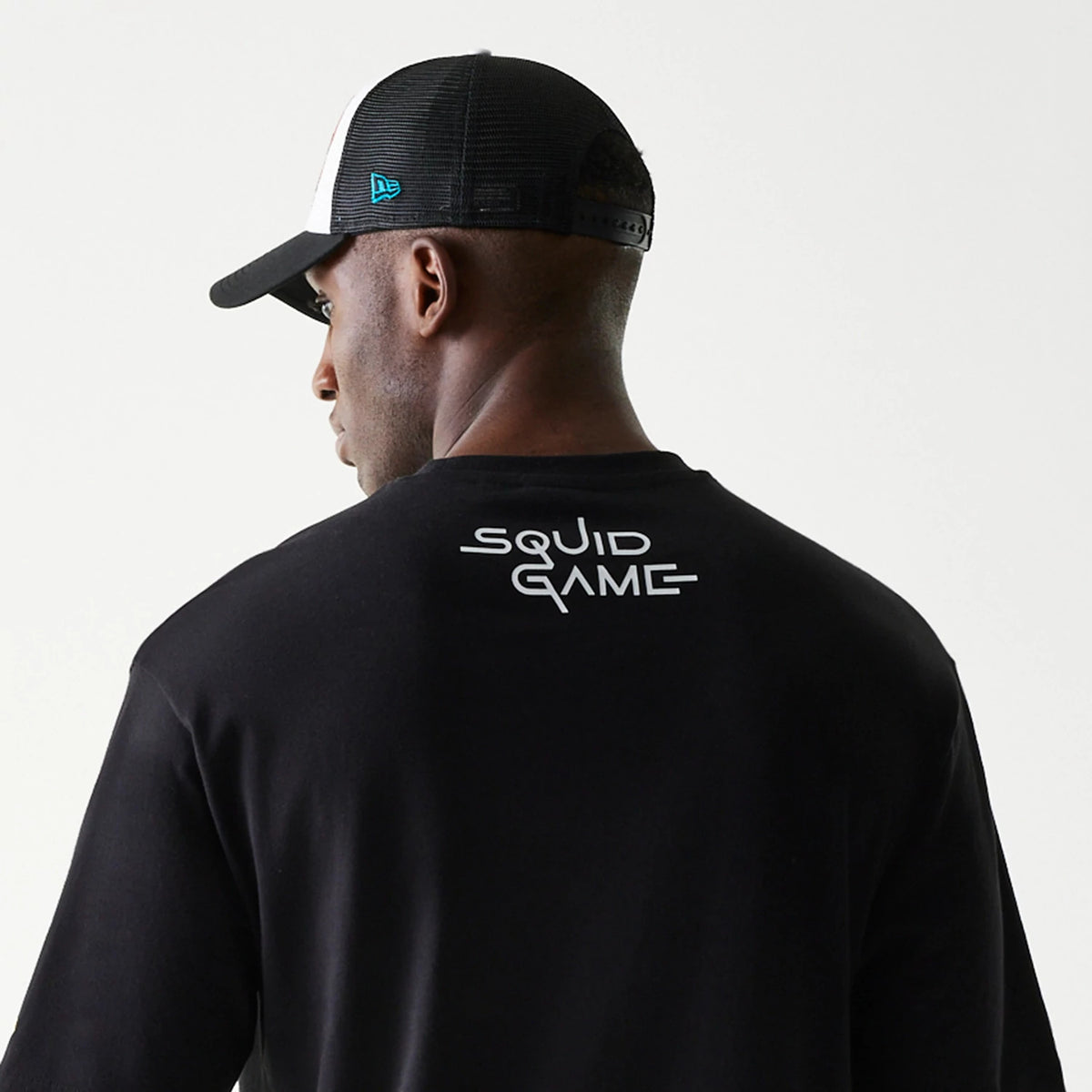 New Era Netflix Squid Game Black Oversized T-Shirt, back view showing the white printed Squid Game logo across the upper back, with a model styled wearing a black and white cap that features a blue embroidered New Era logo on the side.