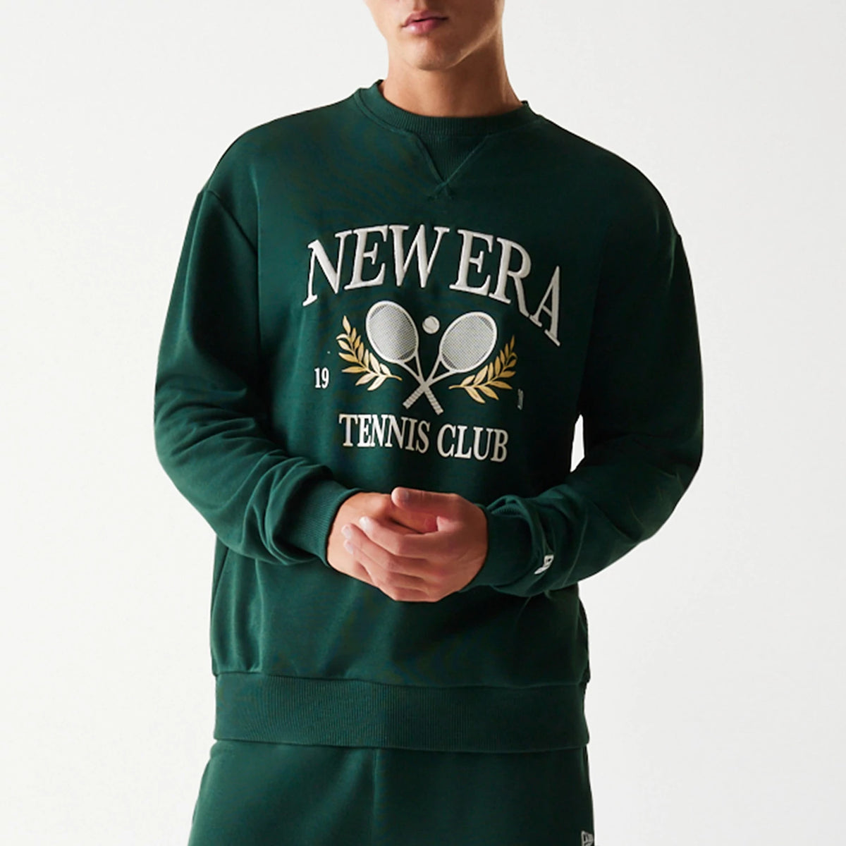 New Era Dark Green Oversized Crew Neck Sweater, front view on a model featuring a large white and gold embroidered tennis club motif with rackets and laurel branches on the chest, ribbed cuffs, and a small embroidered flag logo on the left sleeve.